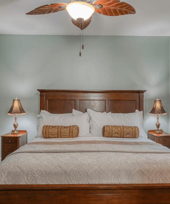 Wood bed, white sheeting, two night stands with lamps, round gold decorative pillows and ceiling fan
