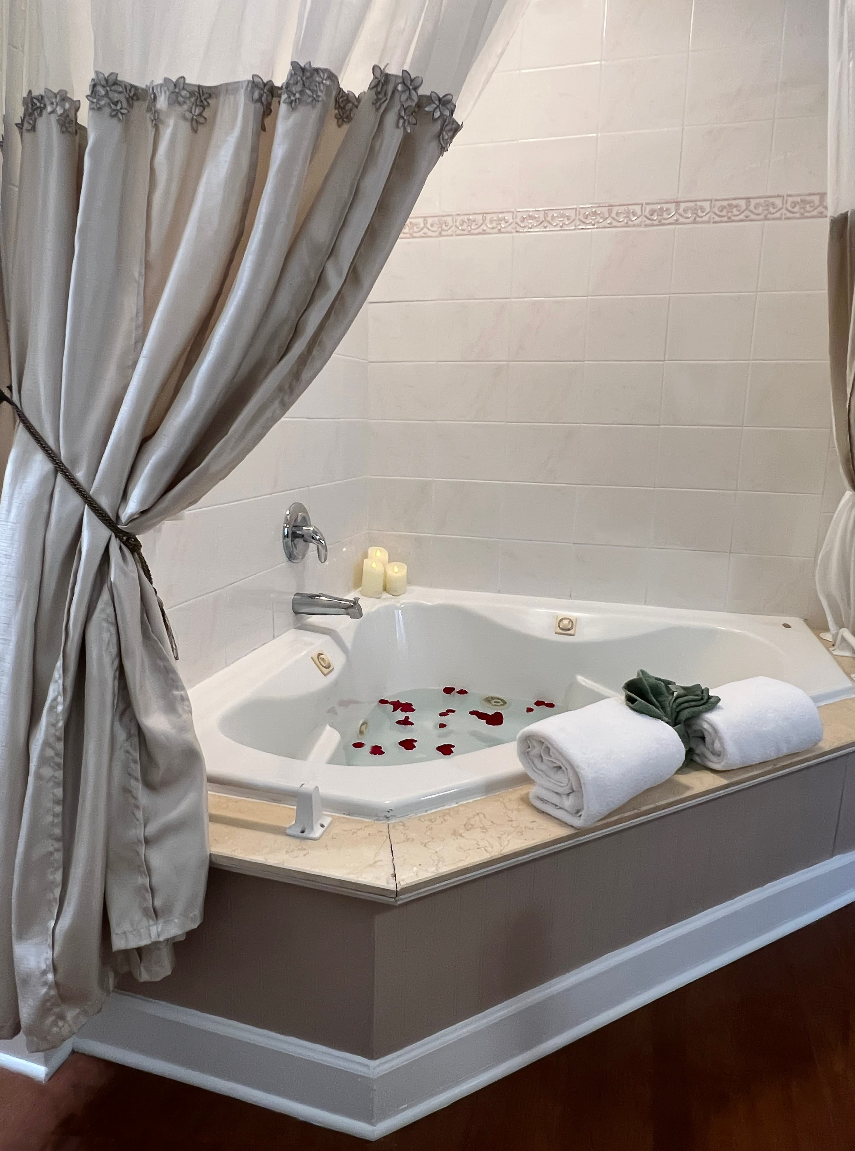 In-room two-person jacuzzi tub with shower, luxuriously draped shower curtain, rose petals floating in water, plush white towels on tub ledge