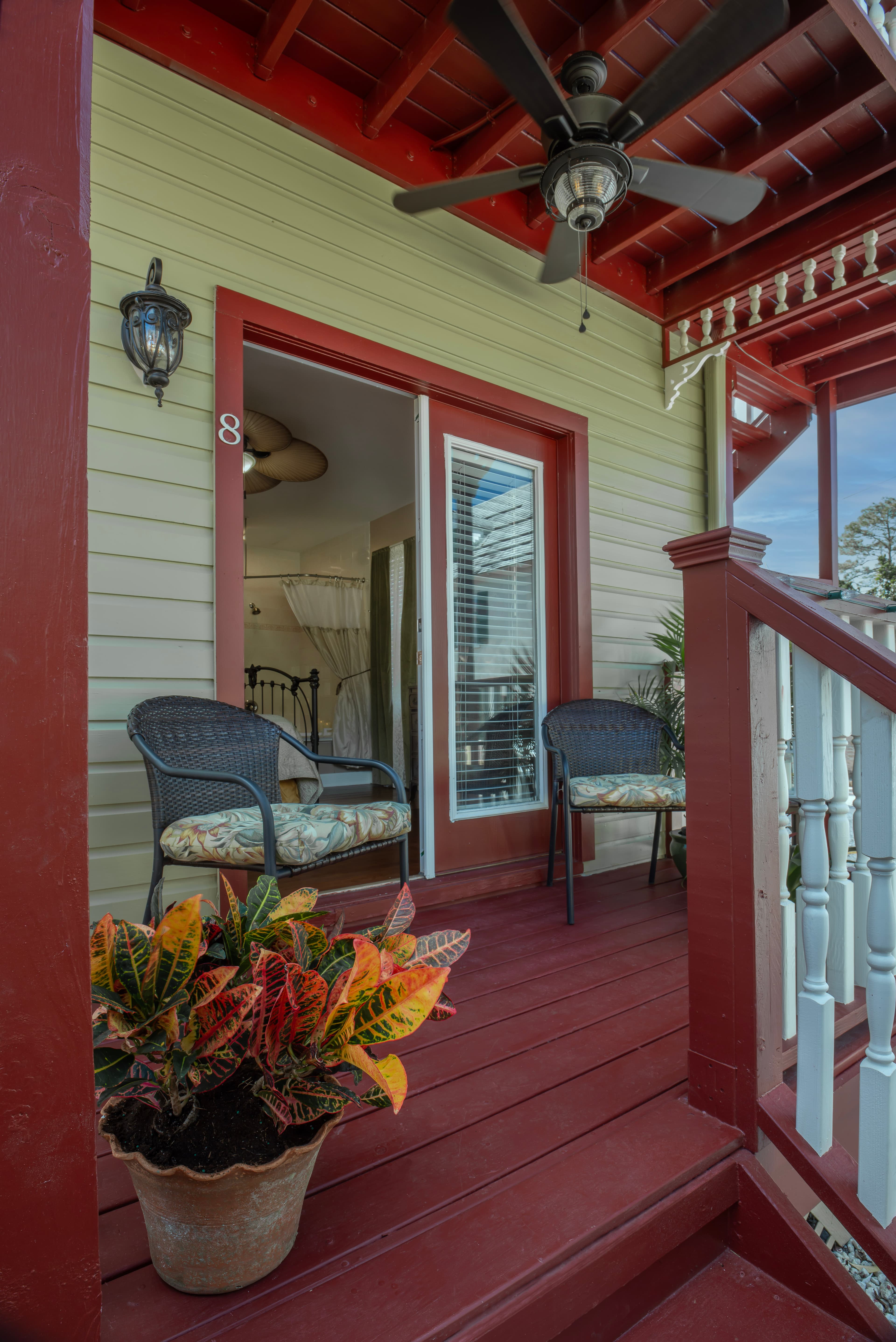 Private porch painted rich red with outdoor chairs, ceiling fan, and potted flowers. Private entry French doors lead to guest room.
