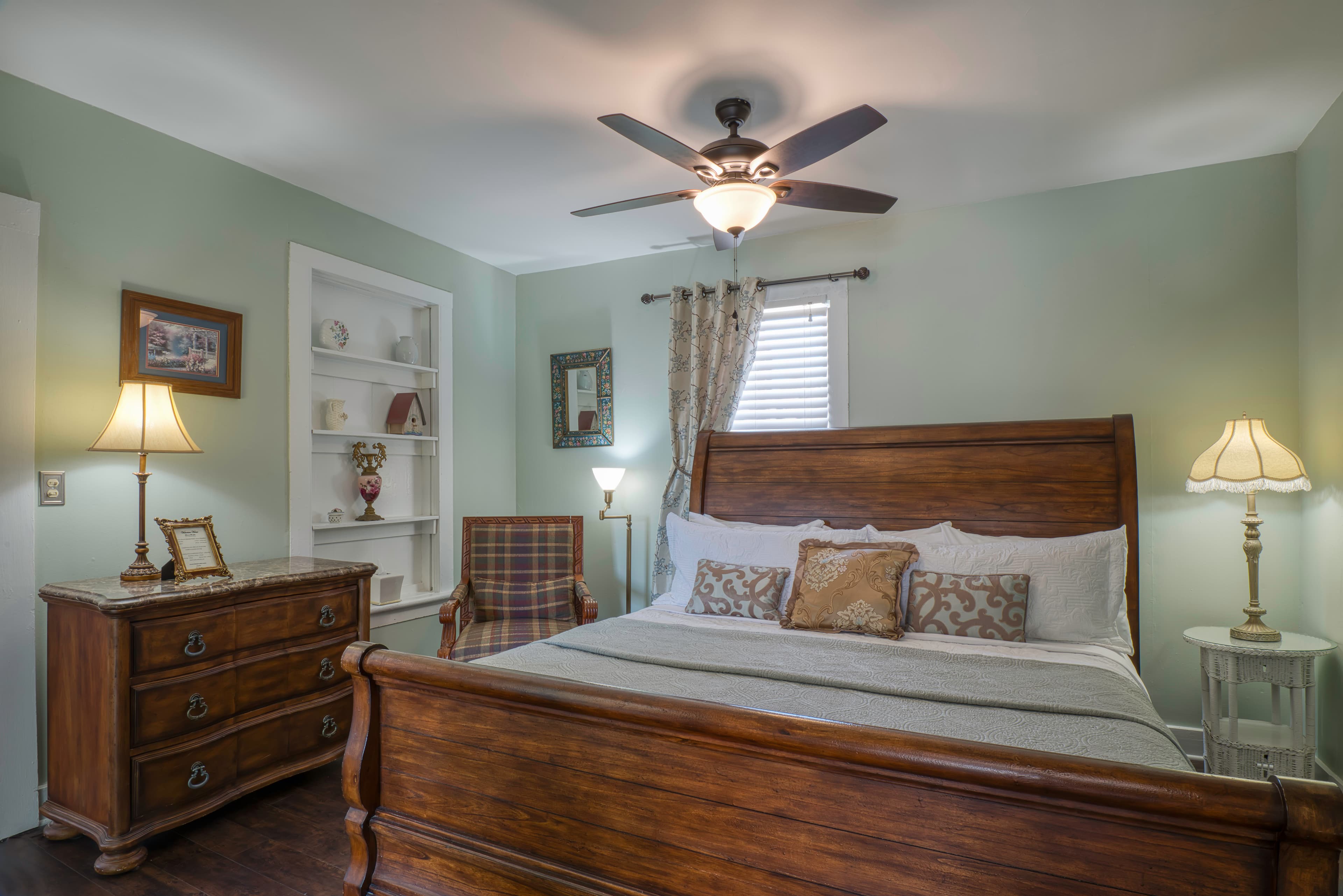 Bedroom in lovely light sage green, king wooden sleigh bed & coordinating dresser, built-in shelves, nightstands with table lamps, and cozy armchair