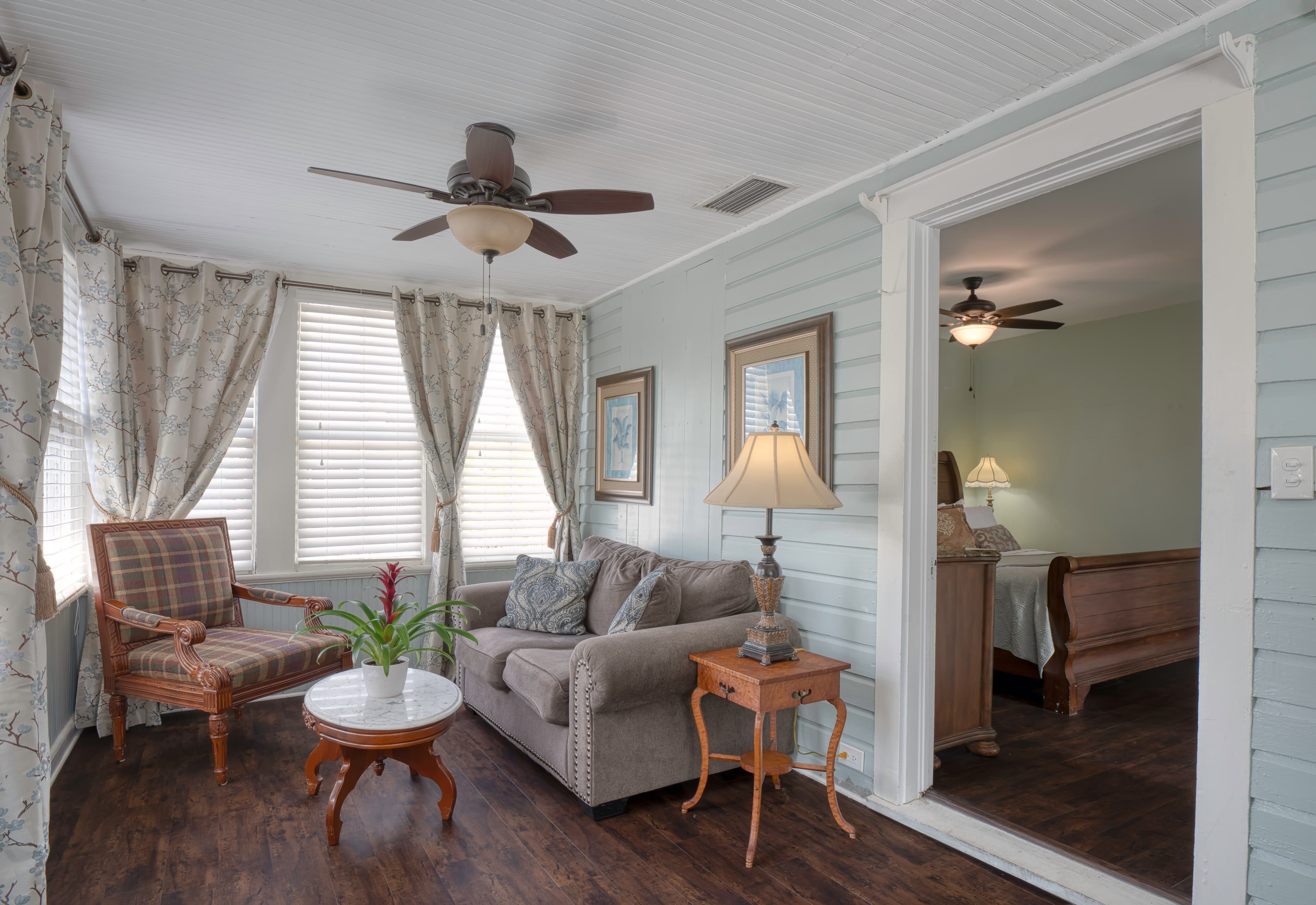 Living room with love seat and armchair, side table with lamp, multiple windows facing Aviles Street, and ceiling fan