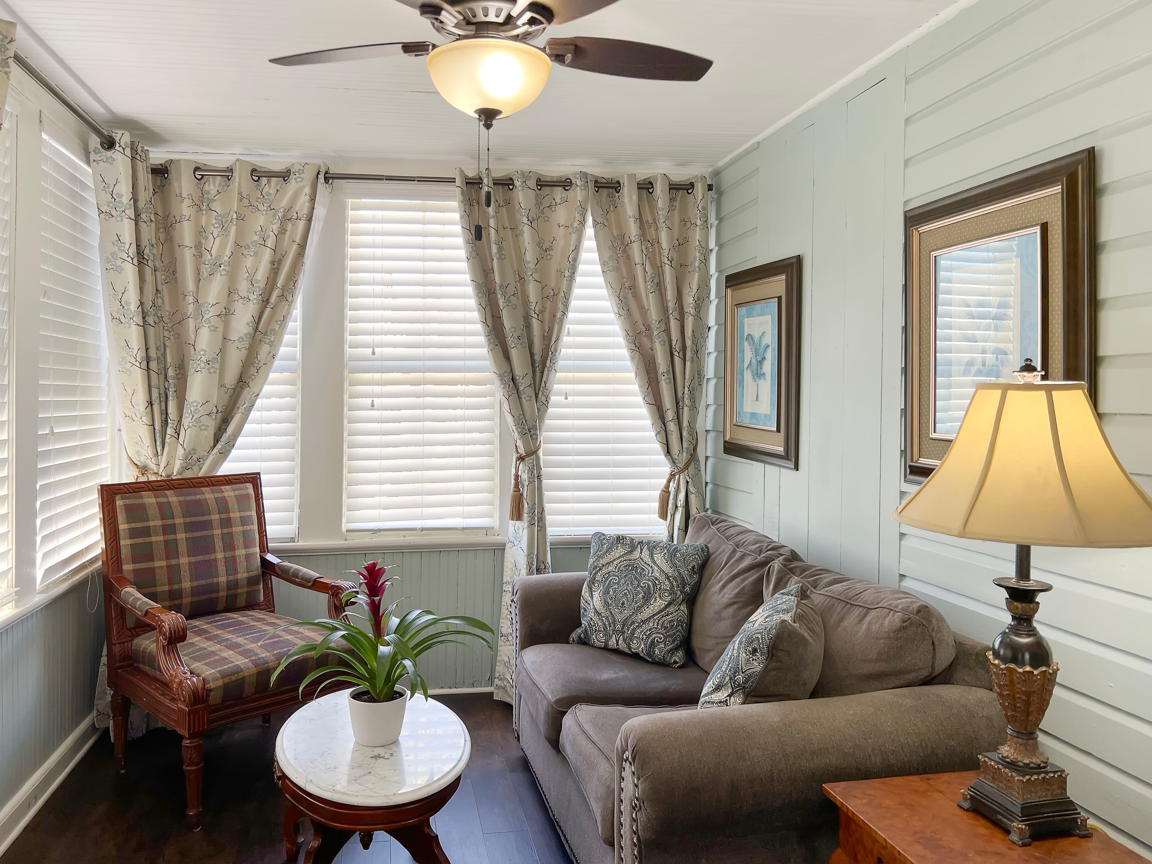 Living room with love seat and armchair, side table with lamp, multiple windows facing Aviles Street, and ceiling fan
