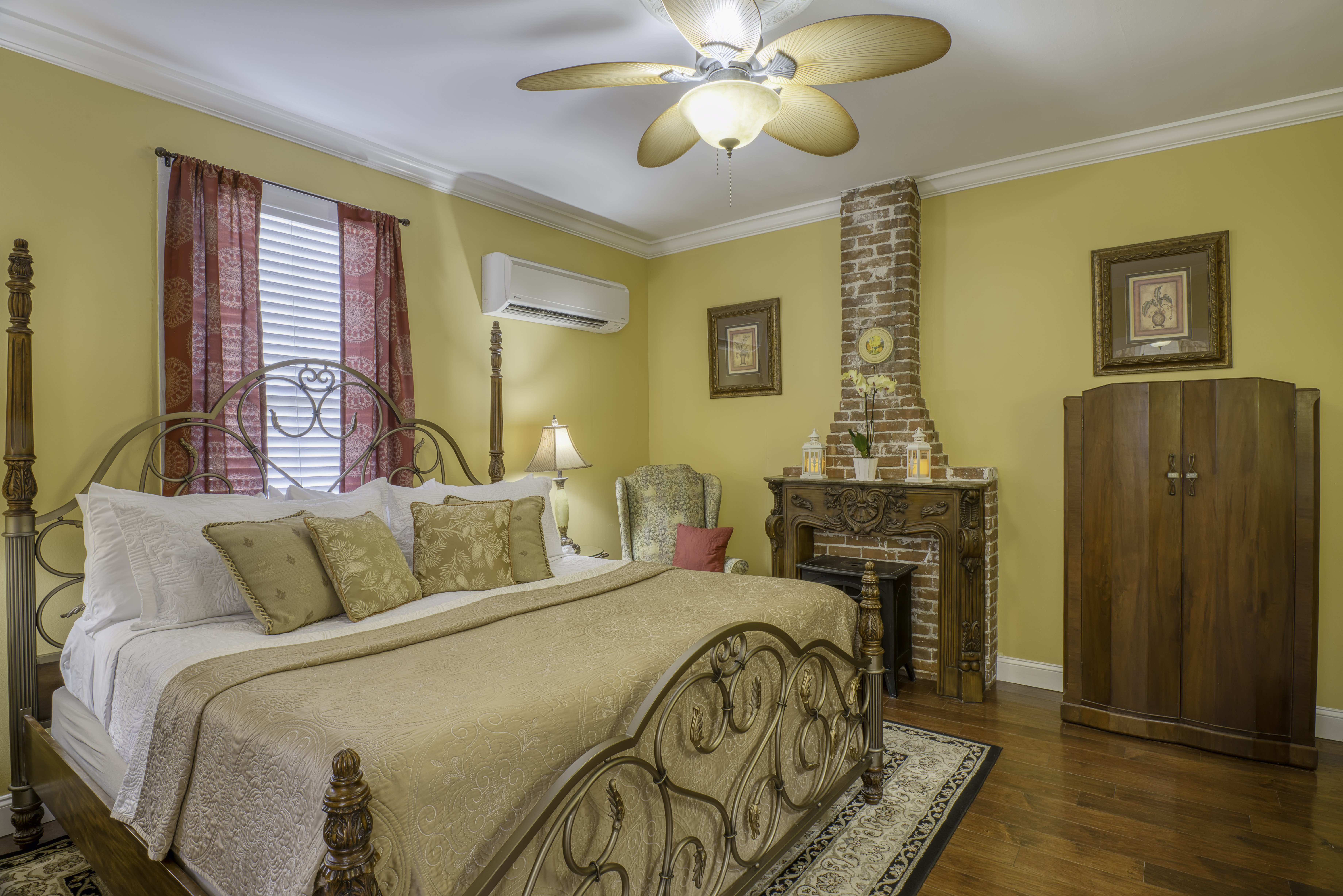 Bedroom in warm yellow and red tones with king brass bed, hardwood floors, electric fireplace, and ceiling fan