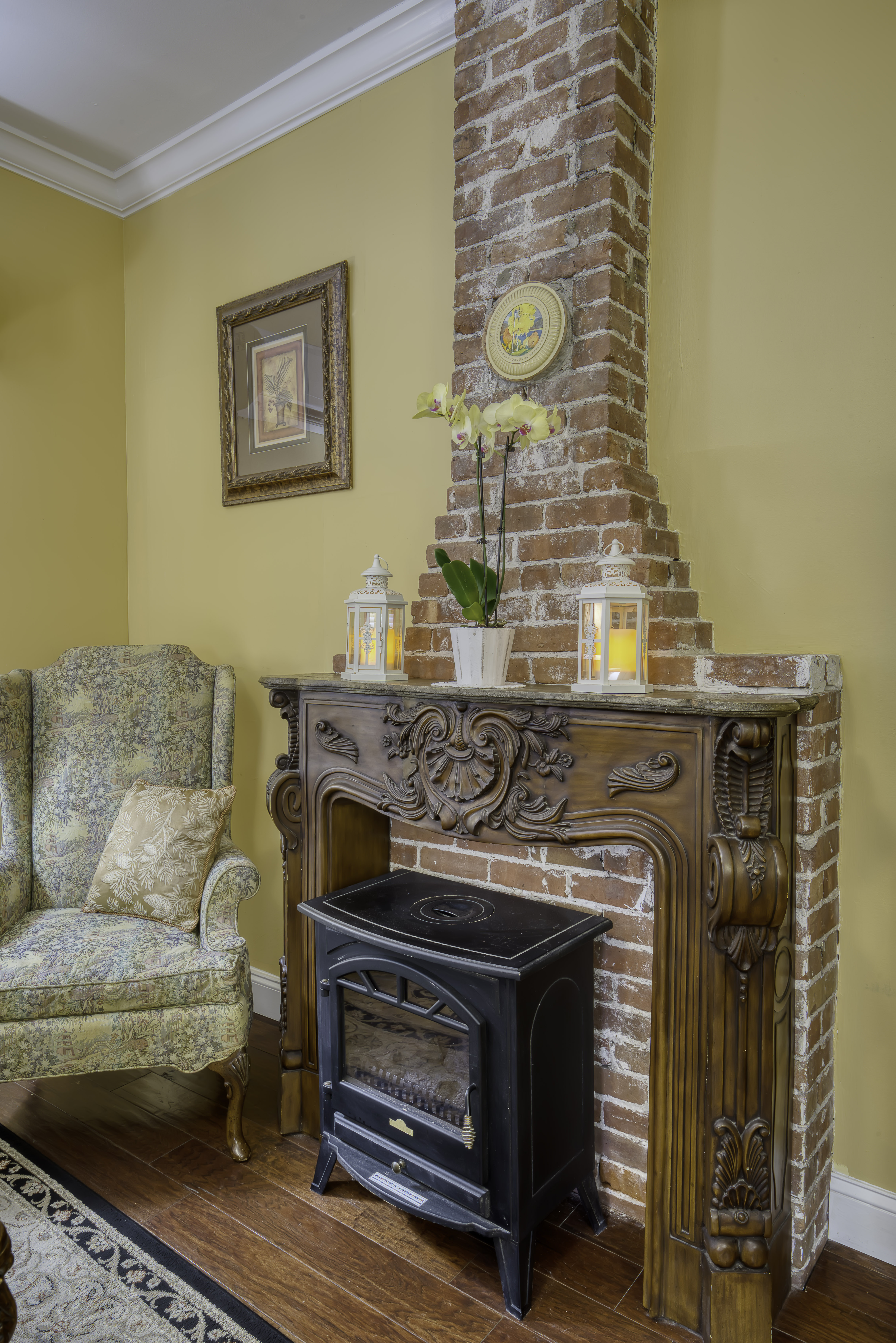 Sitting area with cozy high-back armchair and electric fireplace with brick chimney and ornate wood mantle