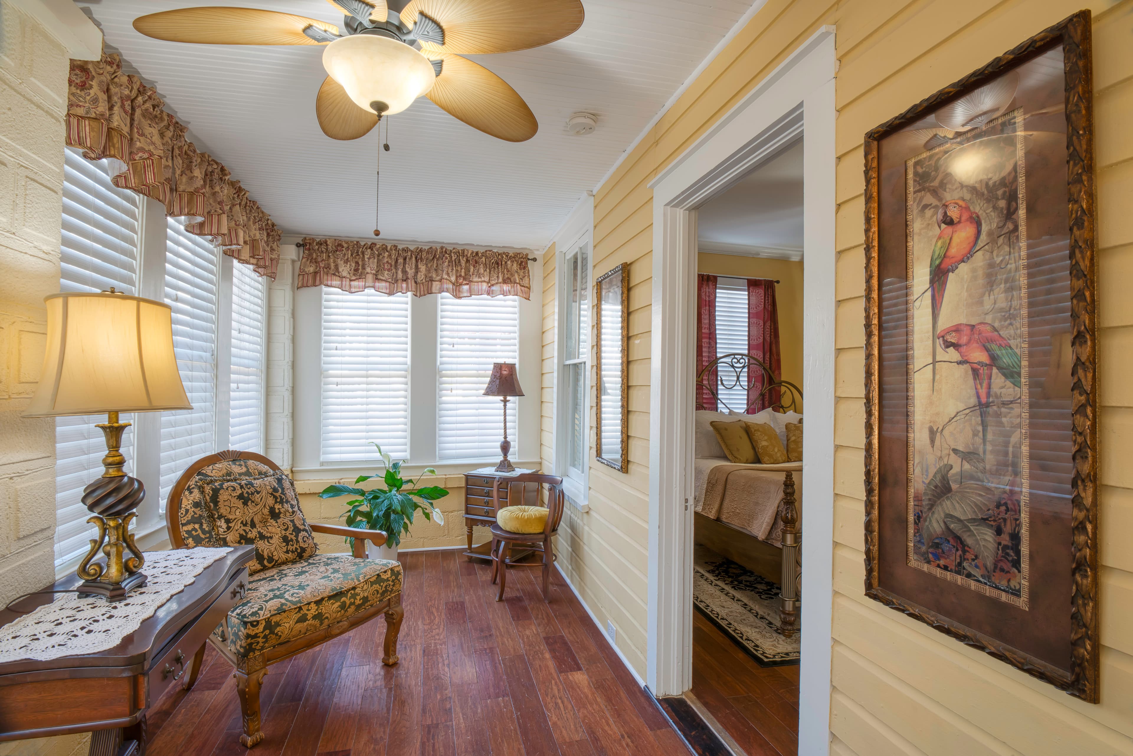 Enclosed private porch with upholstered chair, occasional table with lamp, ceiling fan, and doorway to bedroom