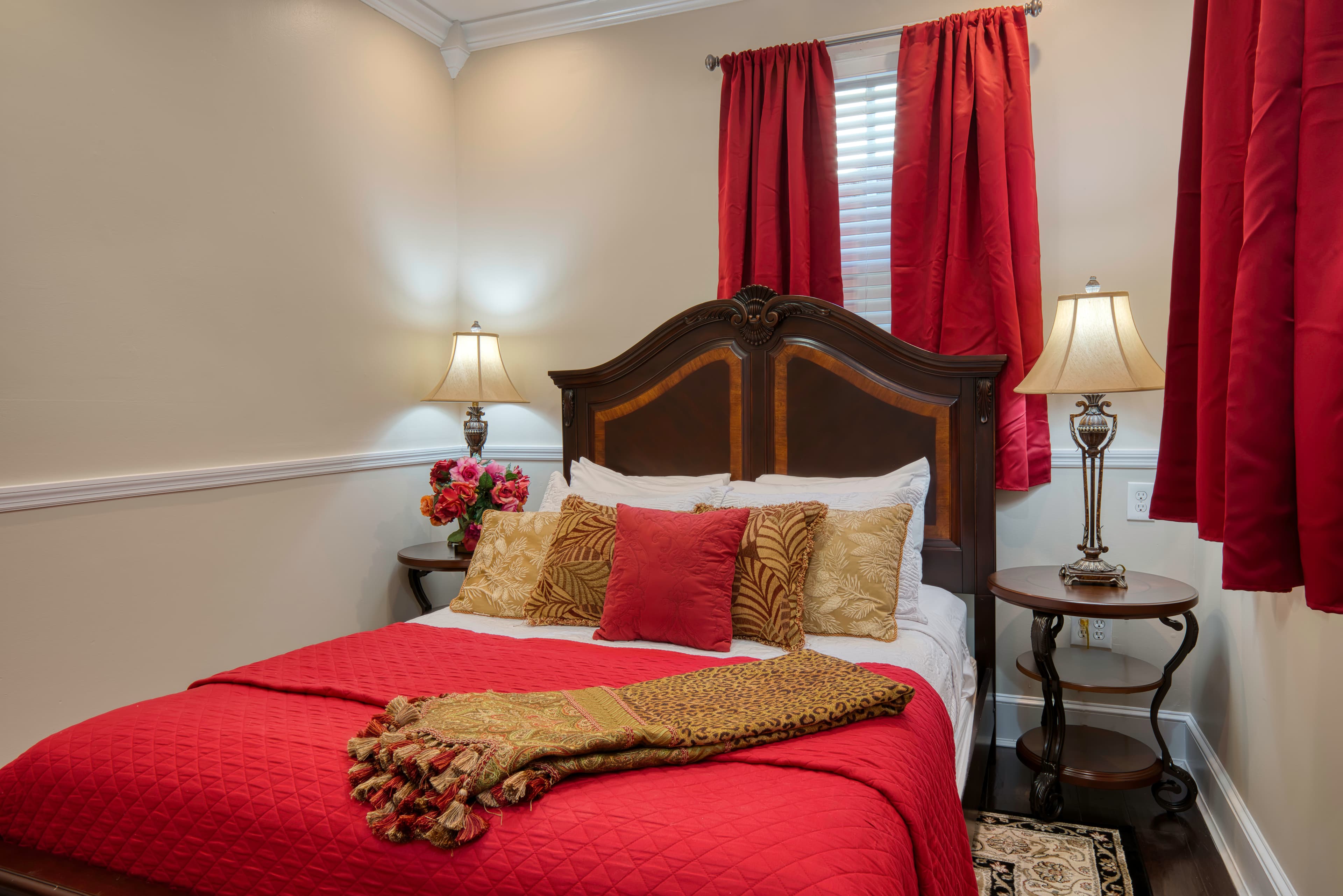 Queen bed with ornate headboard, rich red bedding with gold blanket and decorative pillows, windows with coordinating red curtains, nightstands with table lamps