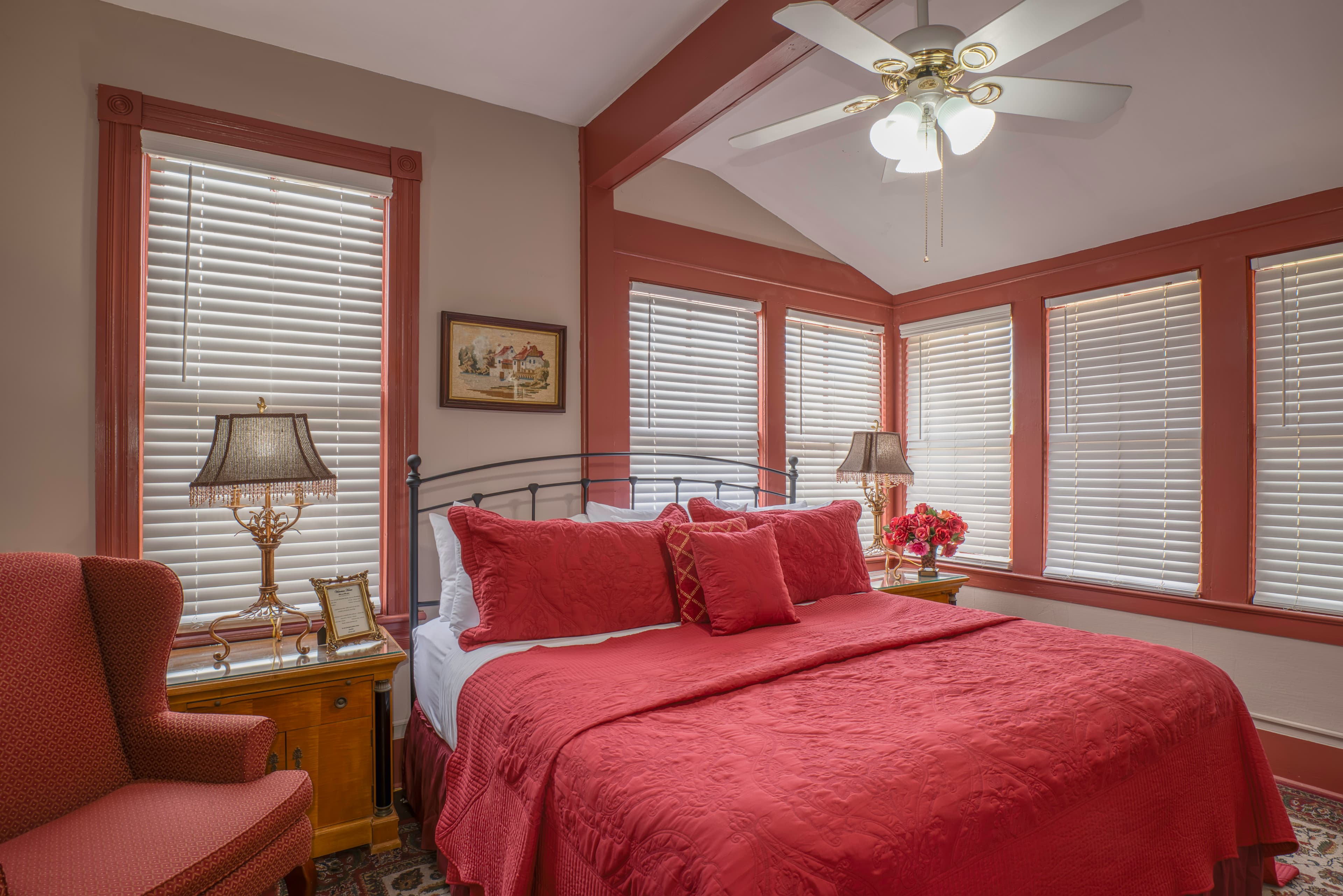 Bedroom with king bed, plentiful windows, matching wooden nightstands with table lamps, comfortable armchair, and ceiling fan