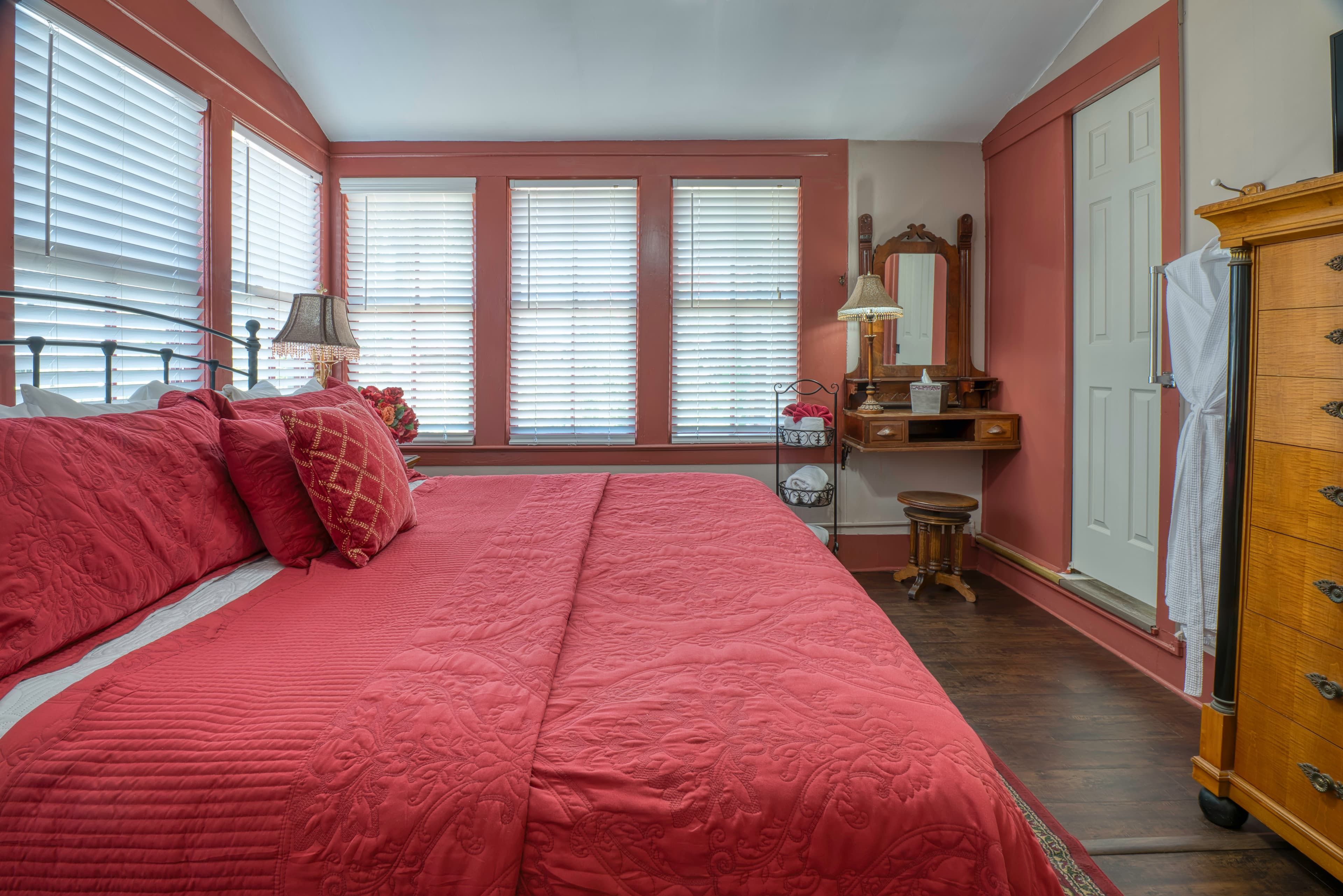 Bedroom with king bed, wooden dresser, vanity with mirror & stool, and sunny windows