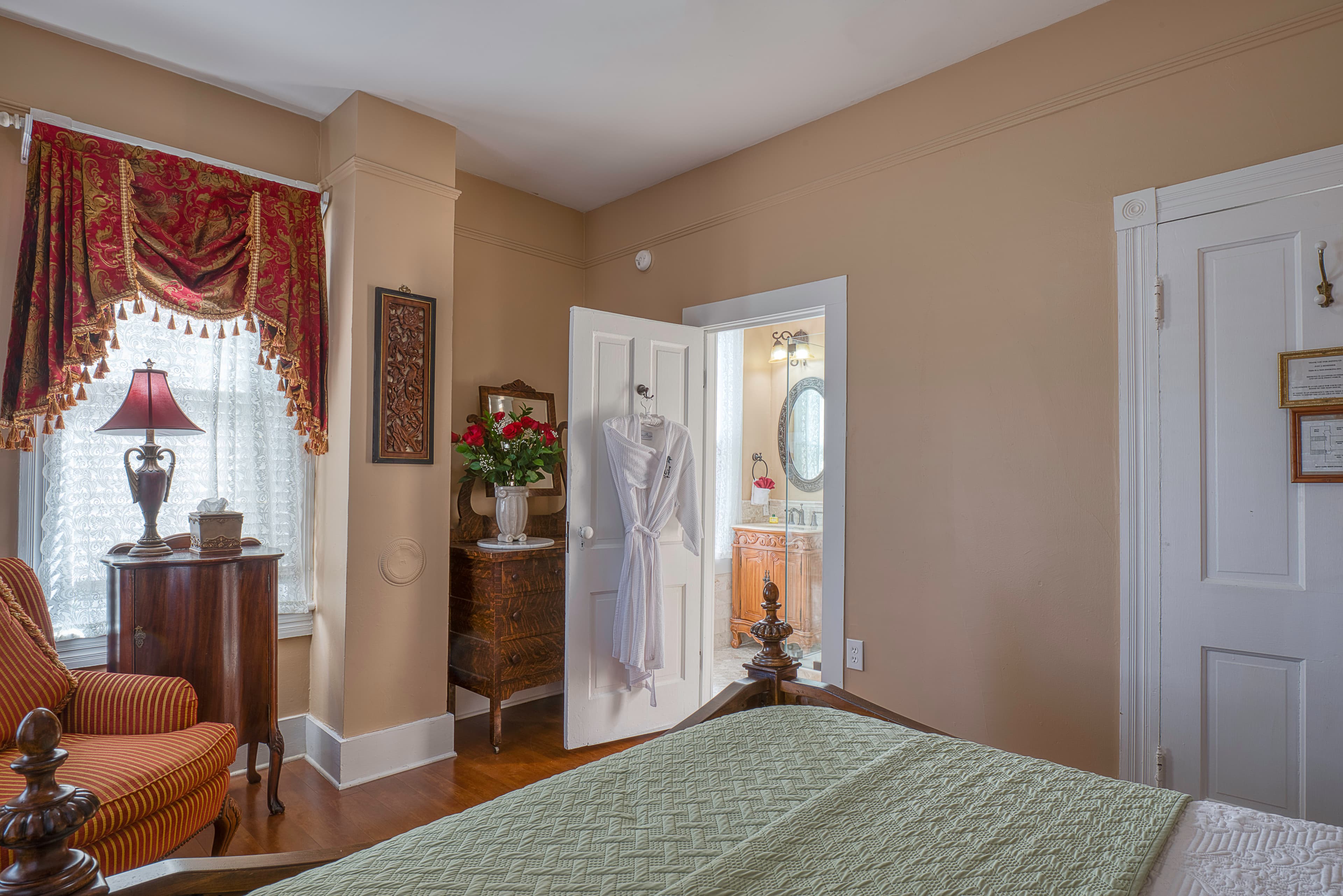 View from bed of sitting area featuring a comfortably upholstered armchair, wooden dresser with flower vase, and doorway leading into bathroom