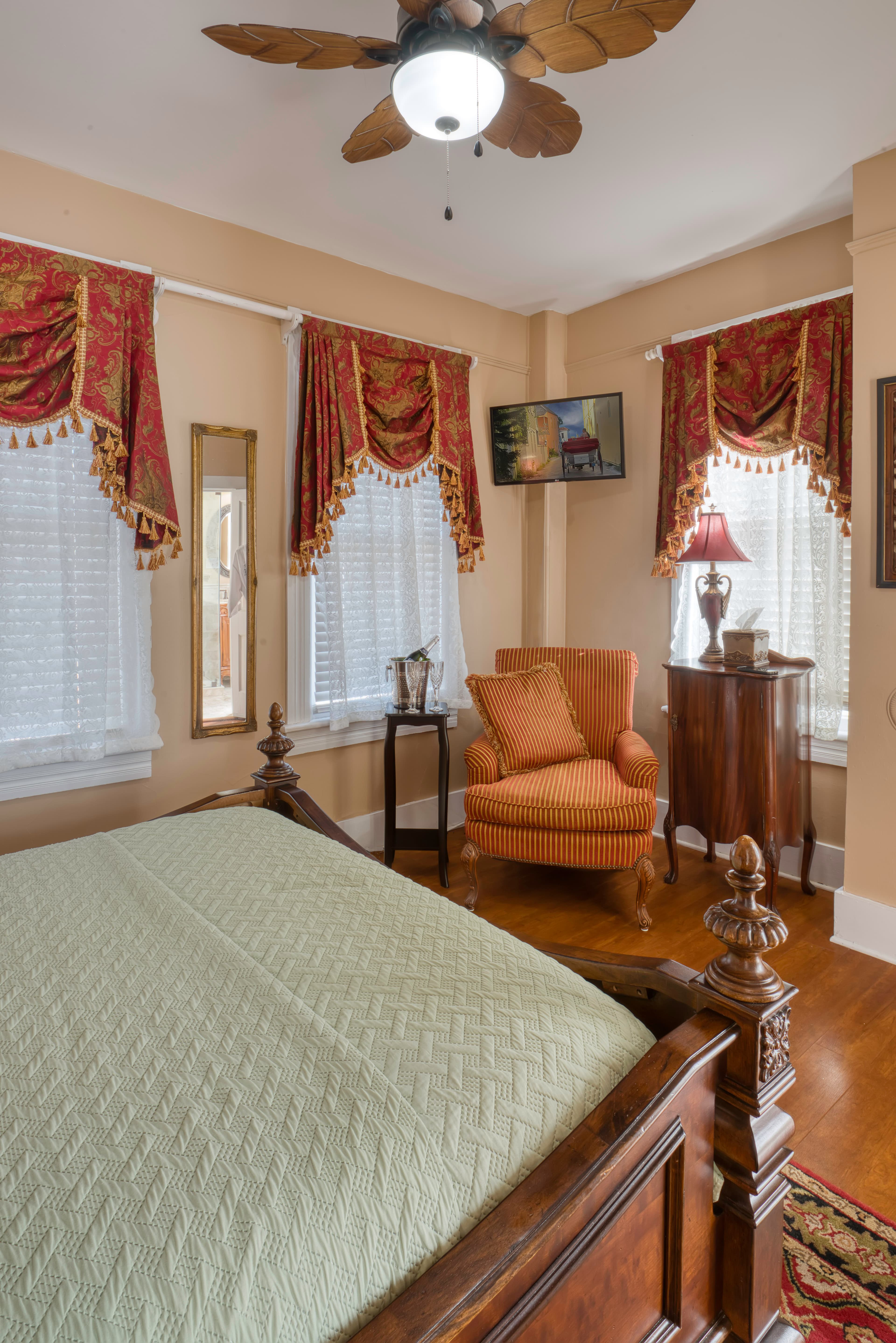 Bedroom with ornate wooden queen bed, hardwood floors, Oriental area rug, large windows opulently dressed in red and gold valances, and a comfortably upholstered armchair