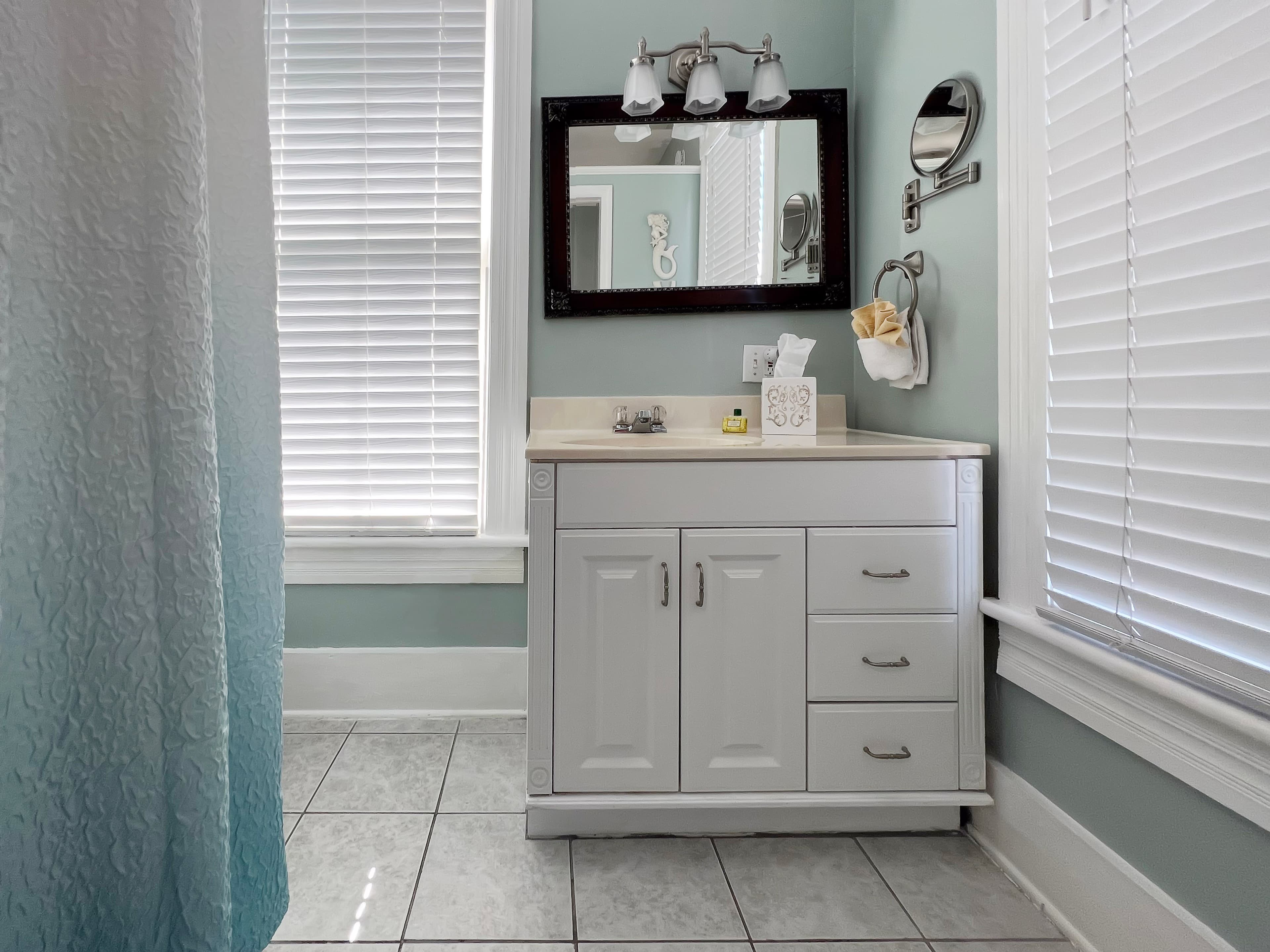 Bathroom in white and shades of blue, white vanity with sink, large windows with blinds,  and shower