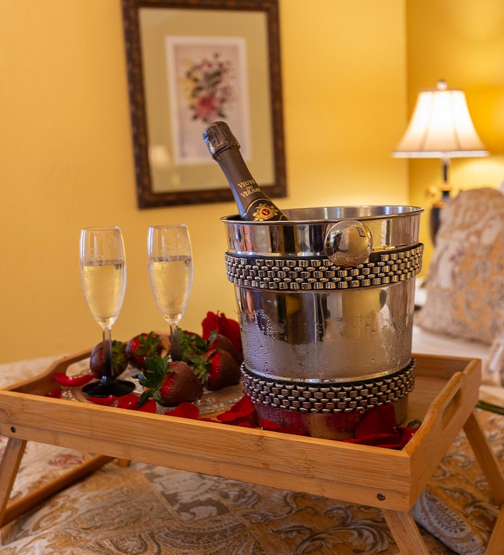 A silver champagne bucket with a bottle, two flutes, strawberries, and rose petals on a wooden tray.