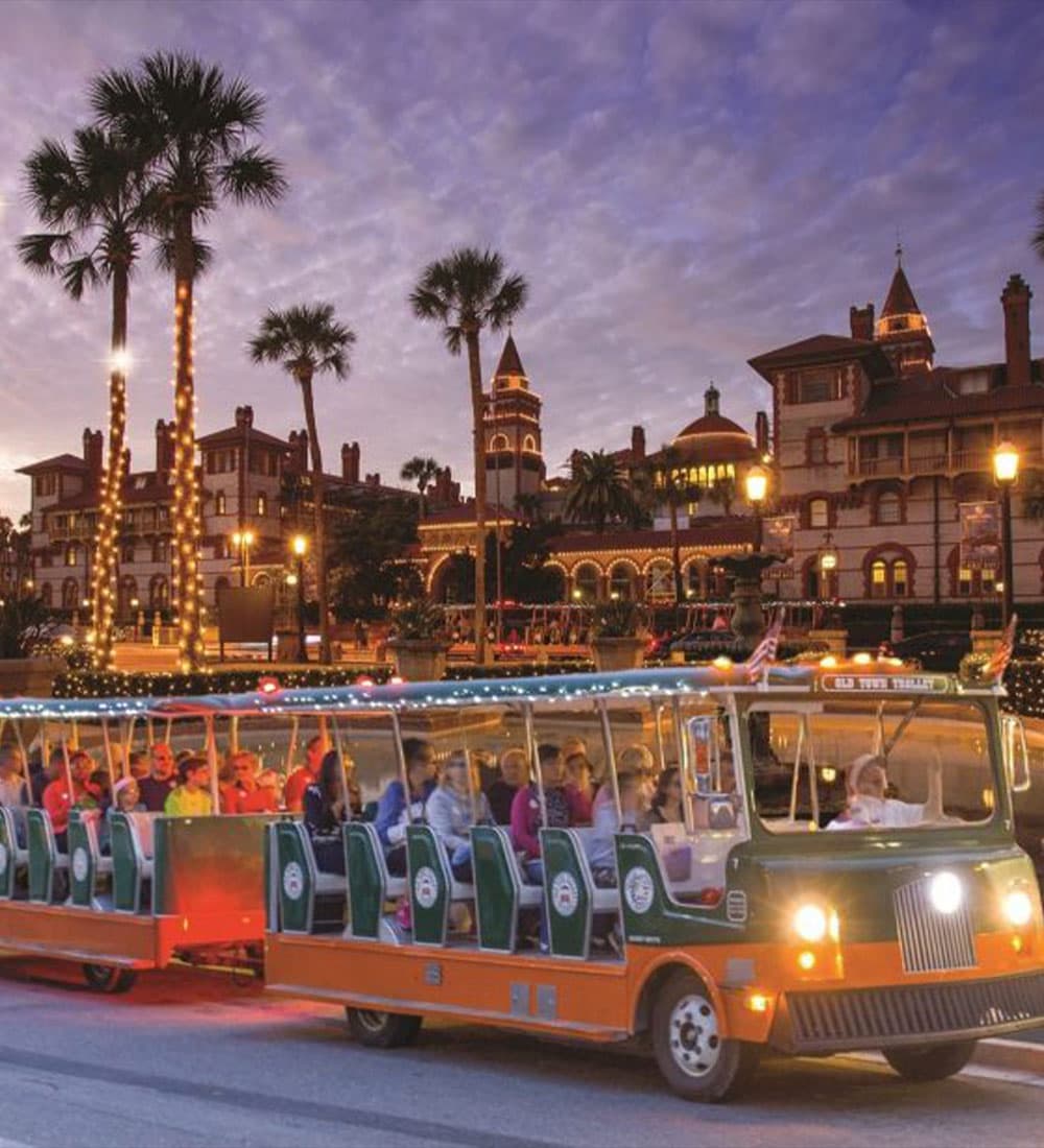 A festive tram filled with passengers rides past a beautifully lit historic building at dusk, surrounded by palm trees.