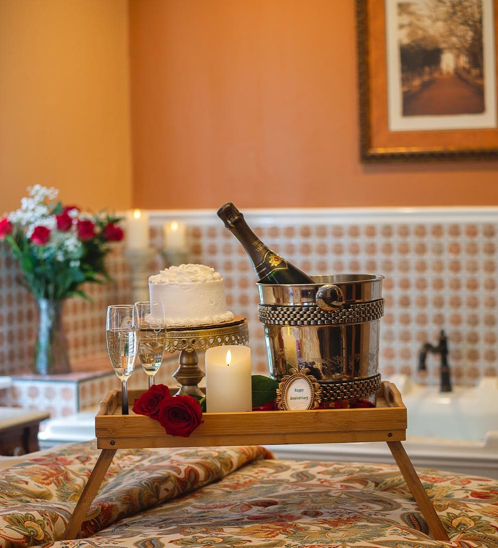 A romantic setup featuring champagne, a cake, and flowers on a tray in a cozy setting.