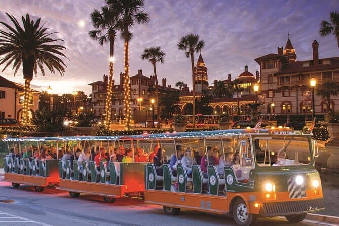 A brightly lit tram filled with passengers rolls past historic buildings and palm trees at sunset.
