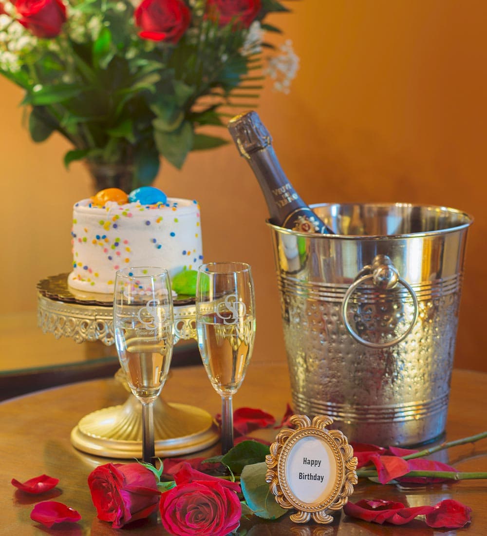 A festive birthday setup featuring a cake, champagne, flutes, red roses, and a "Happy Birthday" sign.