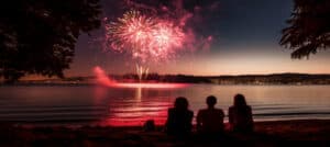 Three people watch colorful fireworks reflecting on the water during twilight.