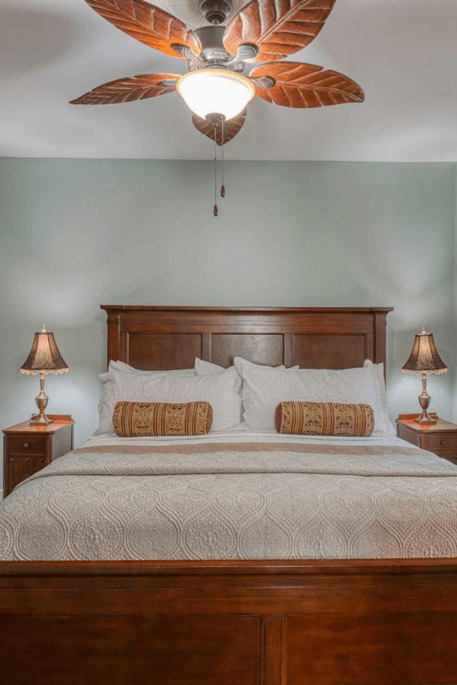 Wood bed, white sheeting, two night stands with lamps, round gold decorative pillows and ceiling fan