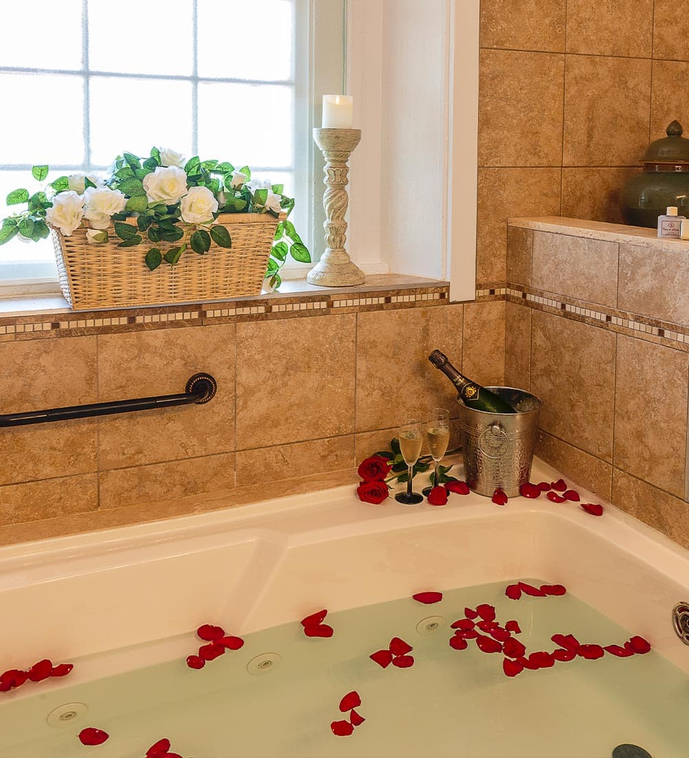 A luxurious bathtub filled with rose petals, accompanied by champagne and flowers on the windowsill.
