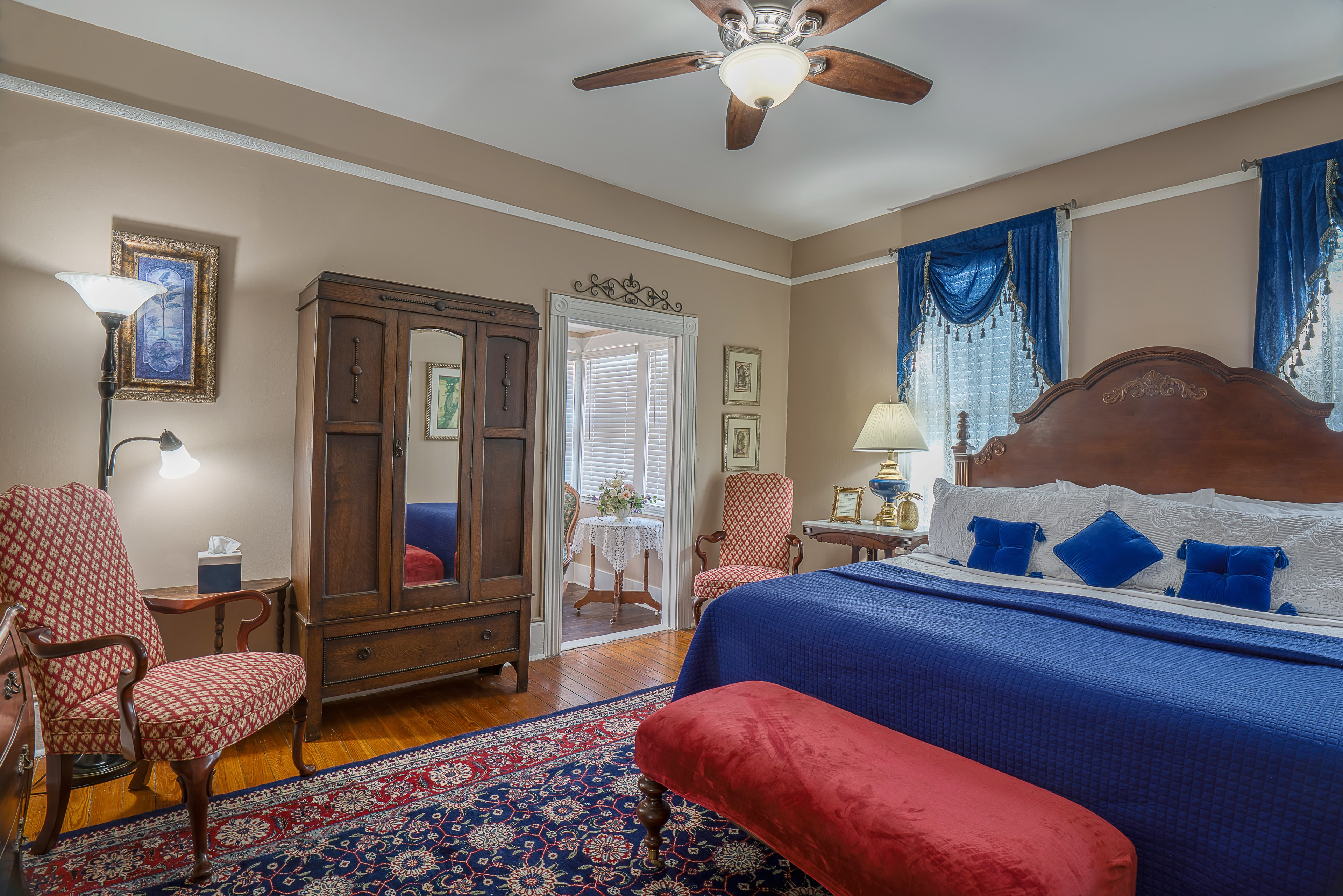 Bedroom with king bed with wooden headboard, coordinating armoire, comfortable armchairs, floral area rug, and upholstered bench at foot of bed