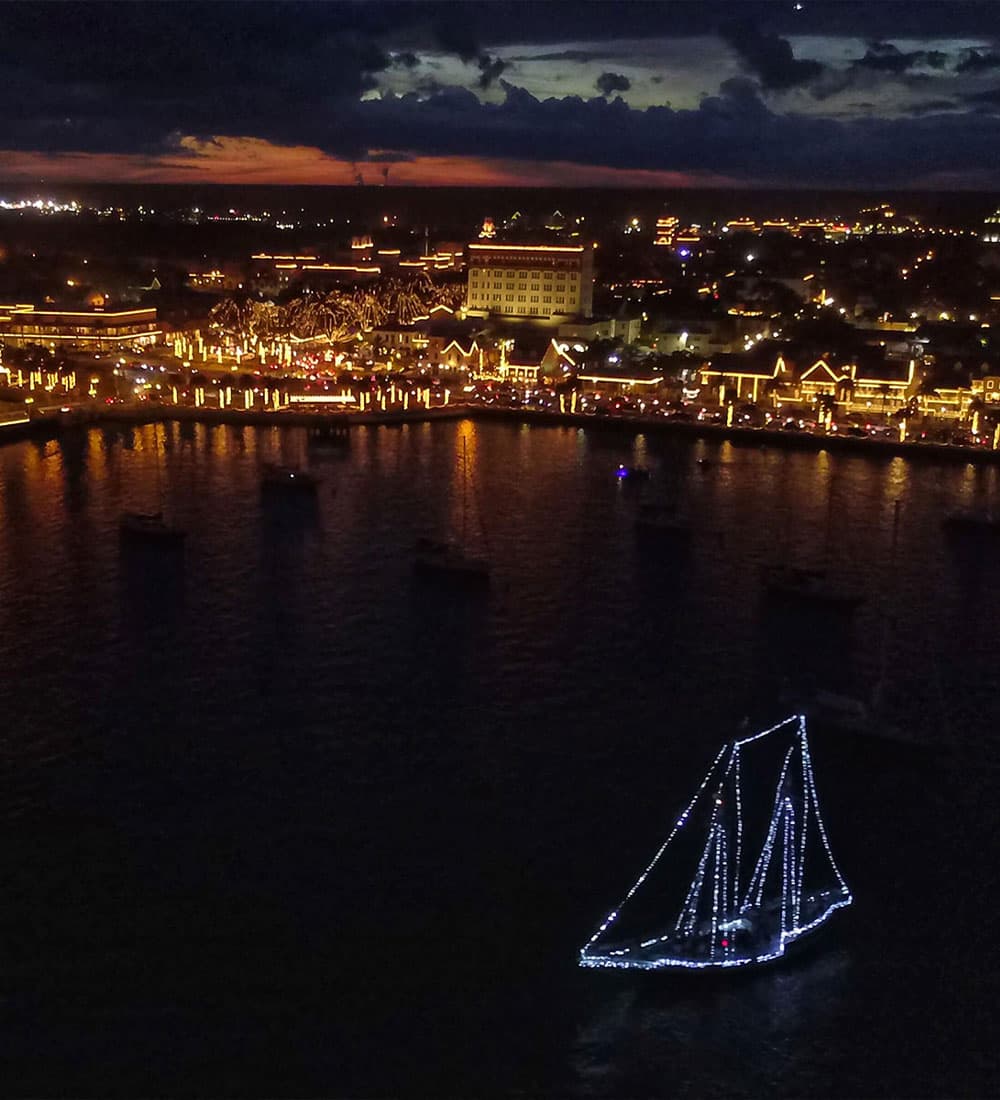 A brightly lit boat sails near a waterfront city adorned with festive lights at dusk.