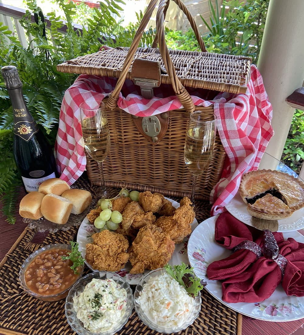 A picnic setup featuring a wicker basket, champagne, fried chicken, grape clusters, coleslaw, baked beans, rolls, and pie on a checkered cloth.