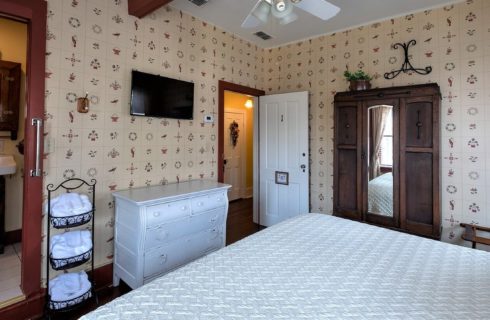 Guestroom with decorative wallpaper, white dresser with TV overhead and doorway into bathroom