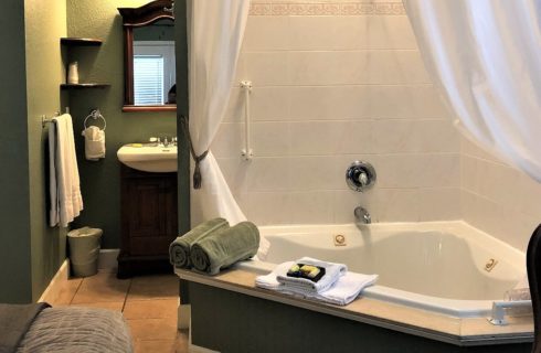 Large corner jacuzzi tub with white curtains in room with bed and single sink vanity