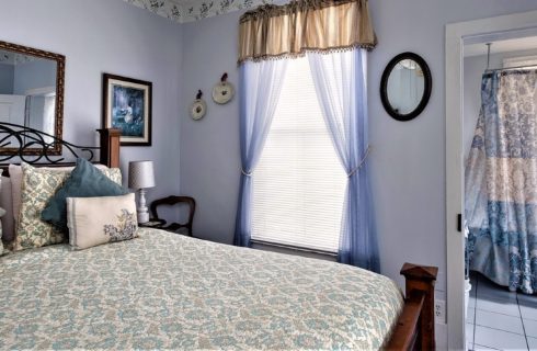 Guestroom with bed in floral linens, large window draped with curtains and doorway into tiled bathroom