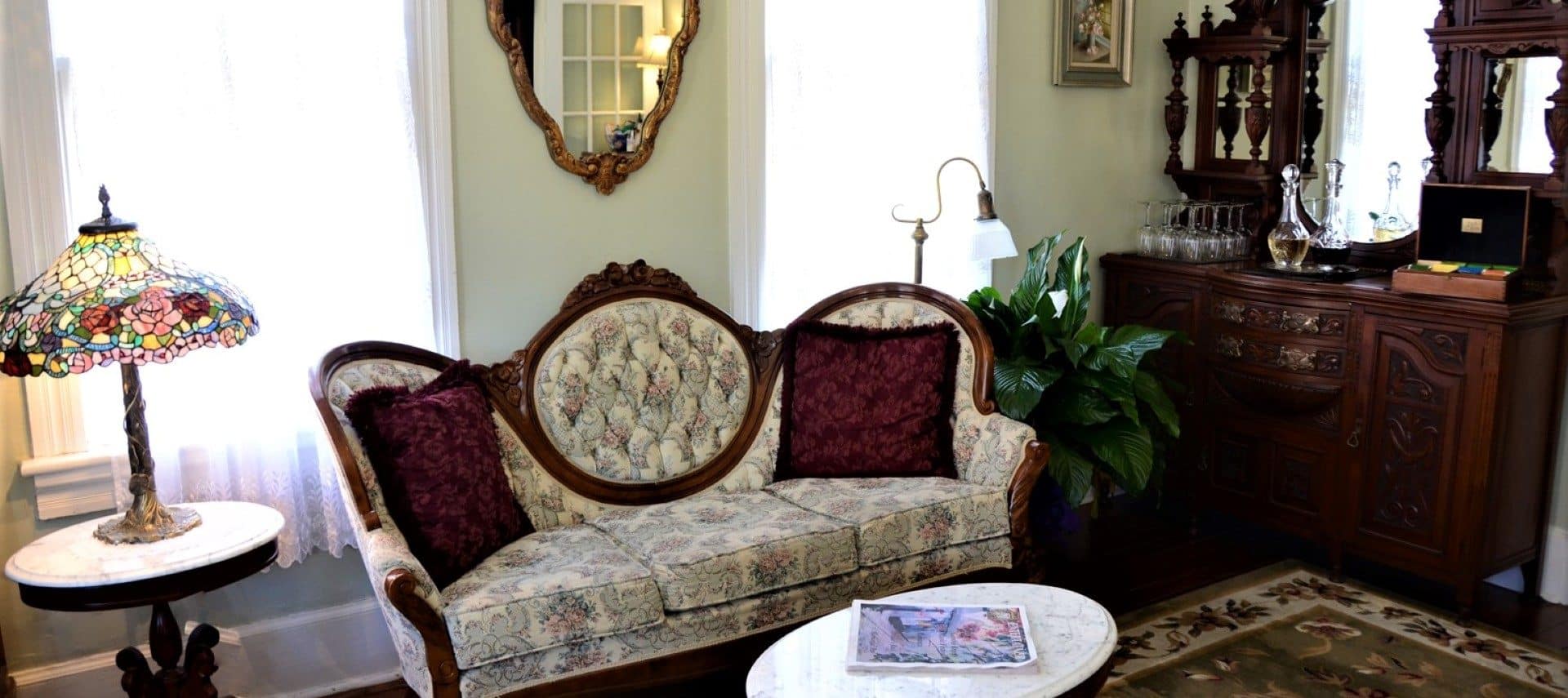 Sitting room with antique floral loveseat, bright windows and side table with Tiffany lamp
