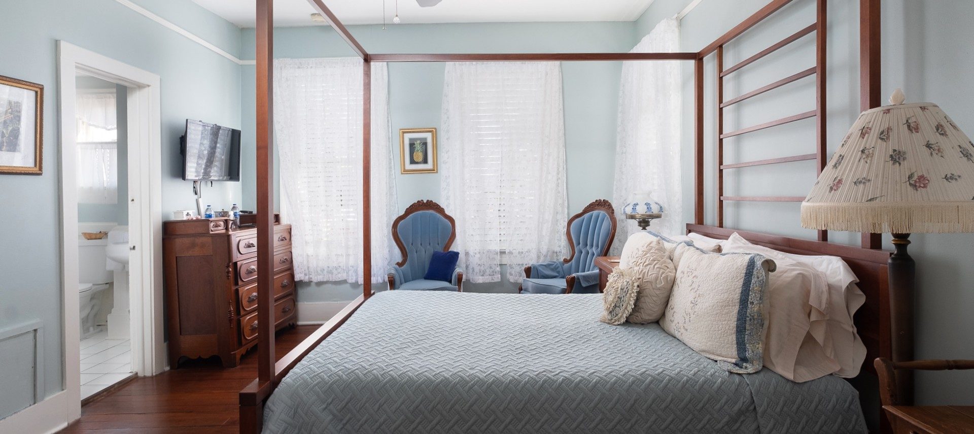 Bright bedroom with four poster bed, two blue sitting chairs, large windows and doorway into a bathroom