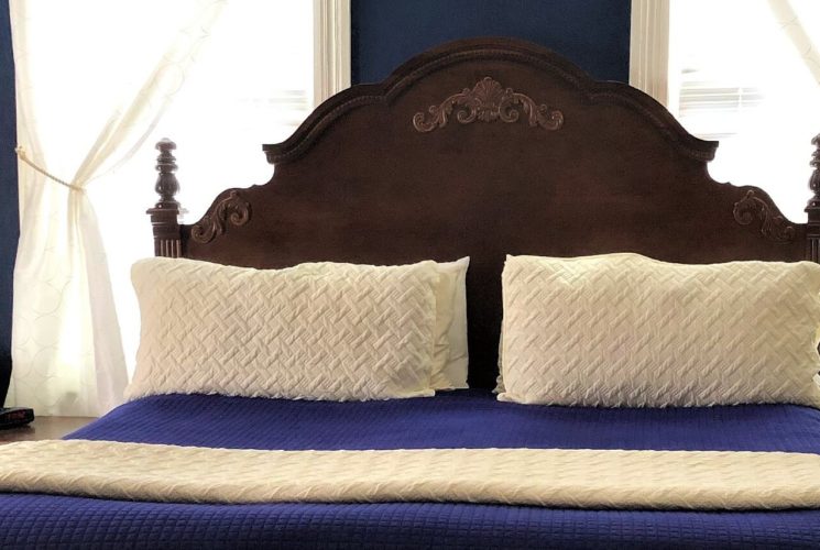 Antique wooden headboard on bed with blue and cream linens in front of large windows, with two sidetables
