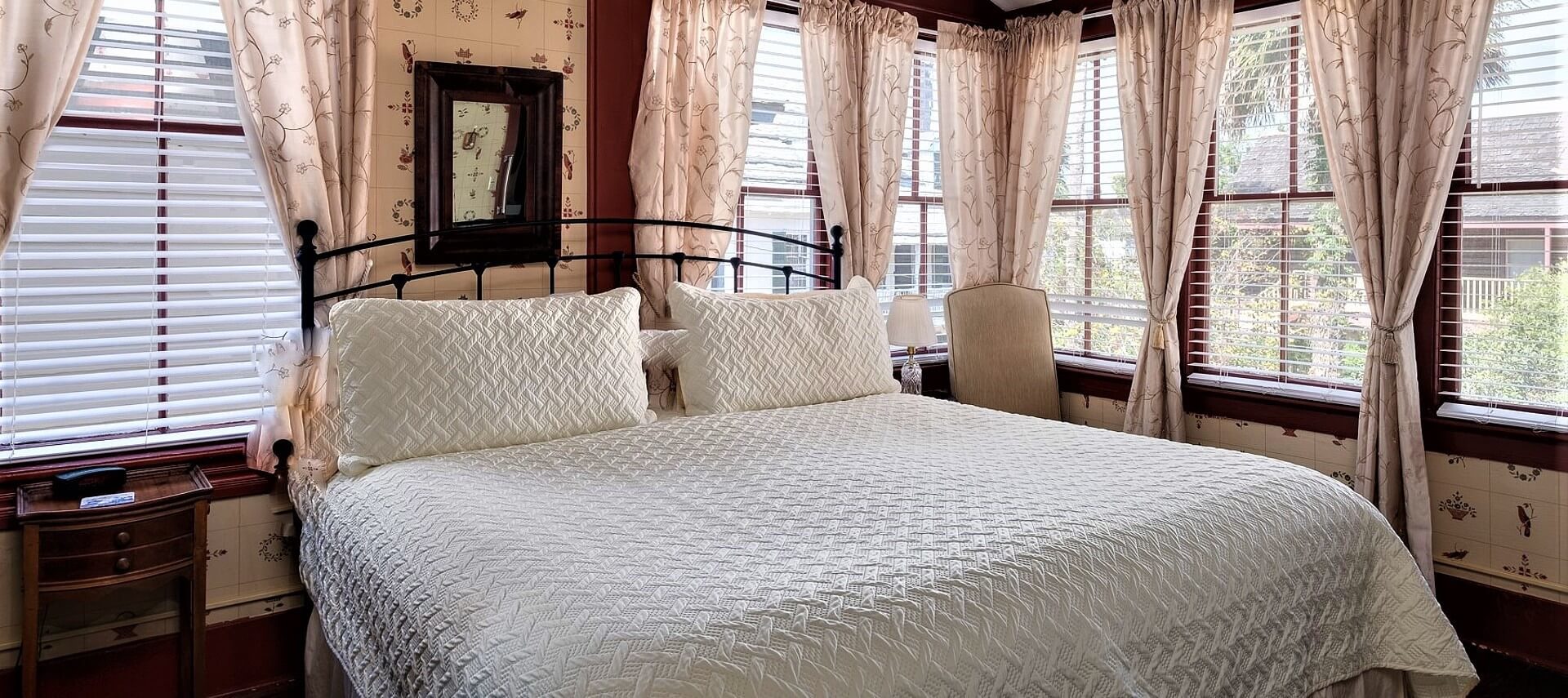 Cozy guestroom with several large windows, bed with cream linens, and wooden side table