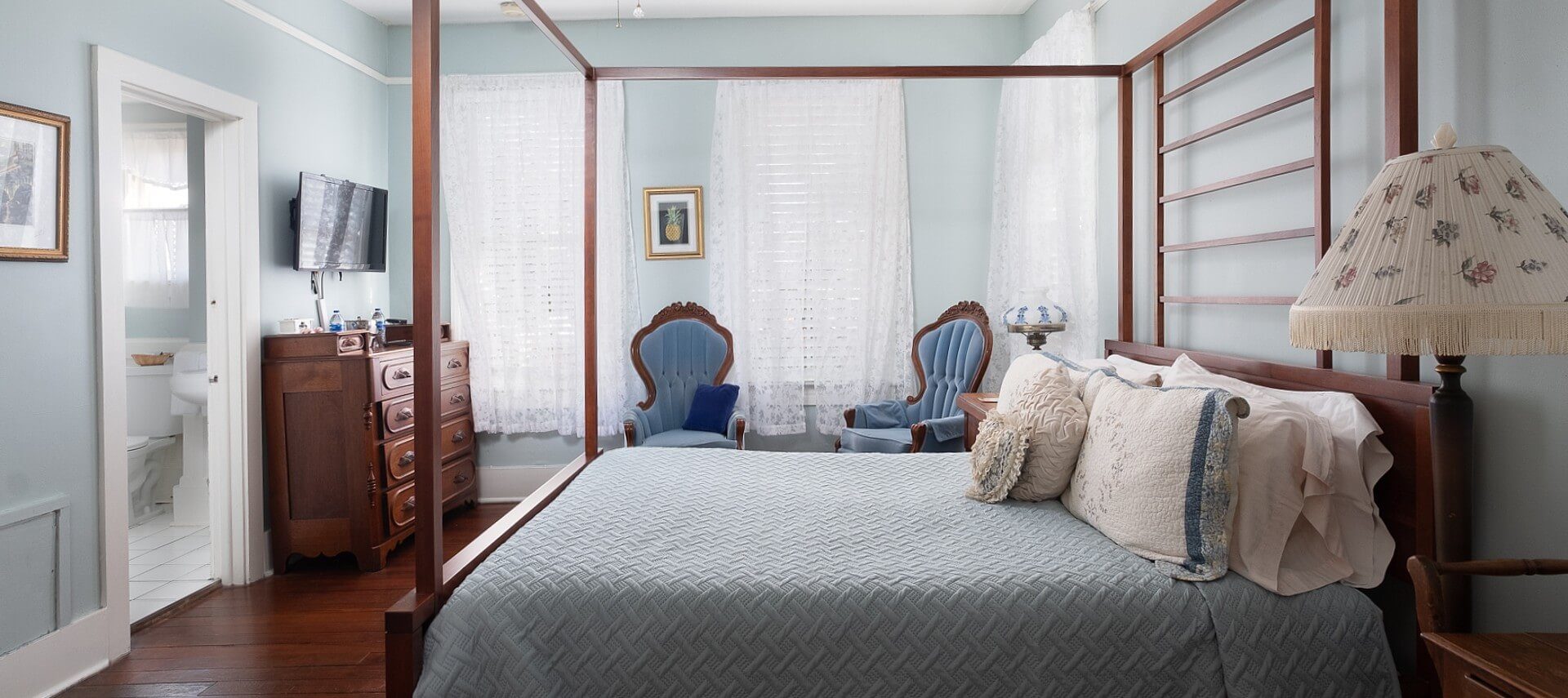 Bright guestroom with four poster bed, upholstered sitting chairs, and wood dresser with TV overhead