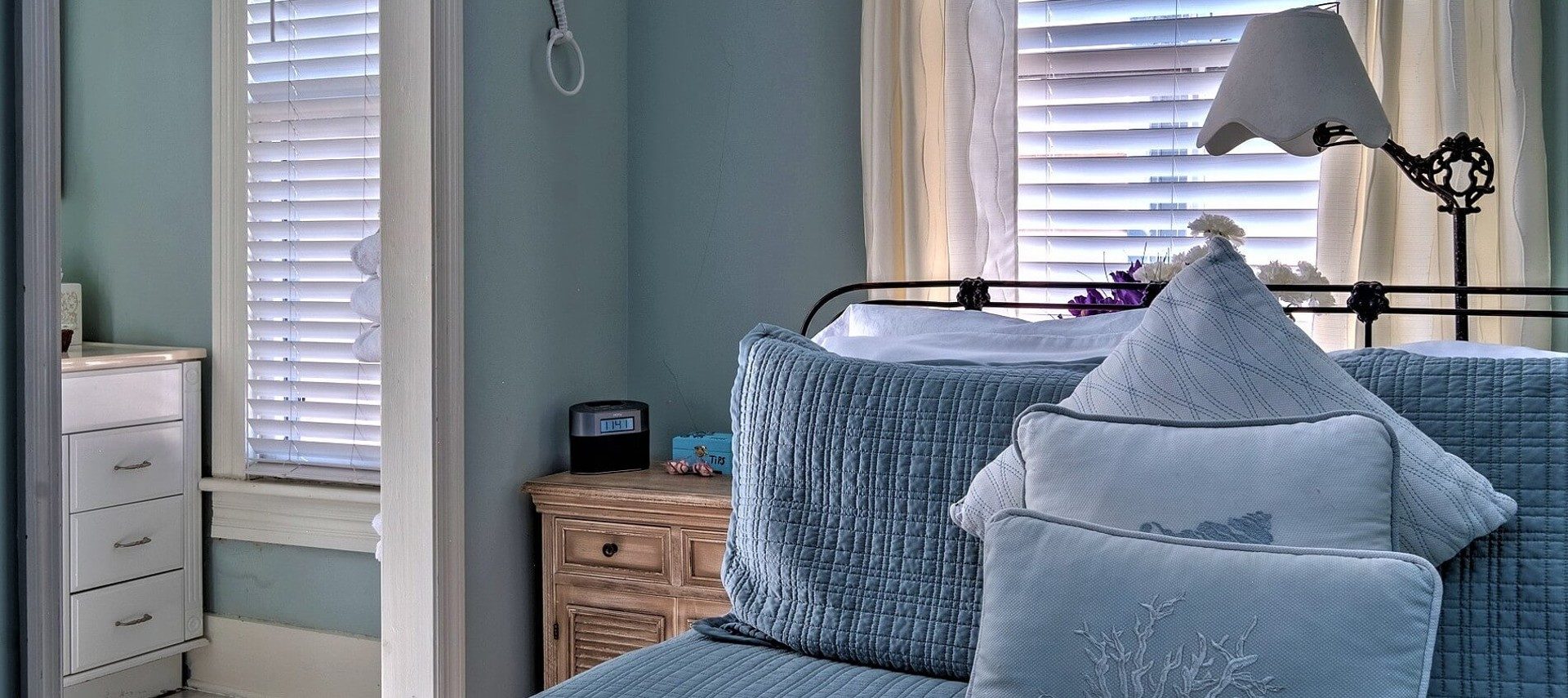 Small guestroom showing wrought iron headboard on bed with blue quilt, window, and doorway into bathroom